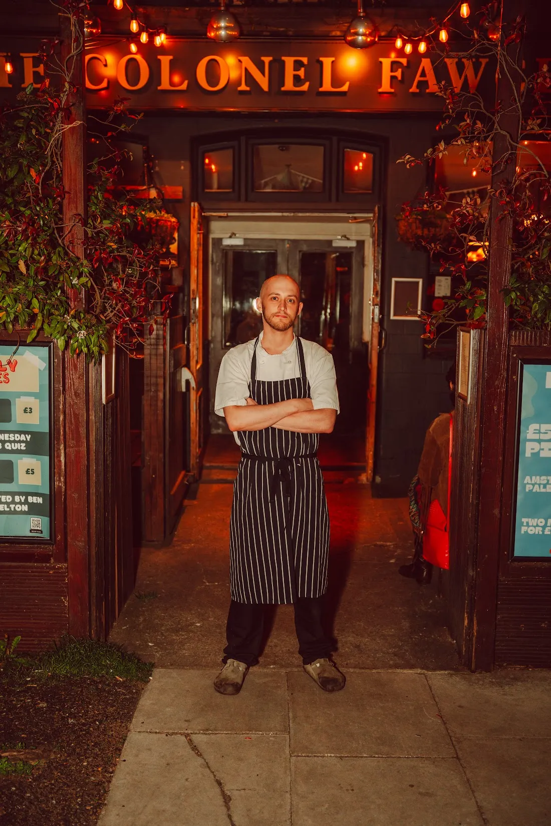 A chef standing alone in the doorway of a pub at night